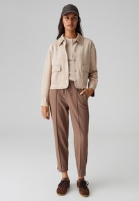 Cappotto in suede beige con tasche frontali, abbinato a pantaloni marroni a sigaretta e sneaker marroni, completato da un berretto scuro.