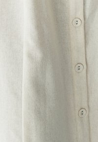 Šviesiai smėlinis lininis marškinis su keturių apvalių mygtukų sagčių eile ir tekstūruota medžiaga. Paprastas dizainas su natūralia išvaizda.