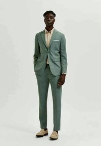 Šviesiai žalia kostiumas pagamintas iš tekstūruoto audinio, turintis vieną mygtuką, liesas kelnes ir baltą kišeninę nosinę, derinamas su tan spalvos marškiniais ir batais.