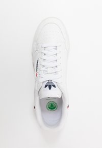 Fehér vegán sneaker peremes orral, lapos fűzőkkel, piros és kék logóval, valamint zöld kerek címkével belül. Lekerekített és texturált talp.