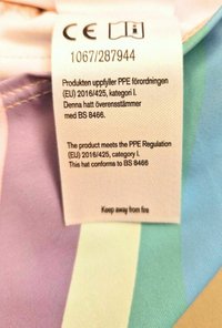 Tarralla monivärisellä kankaalla, jossa on violetteja, vihreitä ja sinisiä raitoja; sisältää hoito-ohjeet ja sertifiointitiedot.