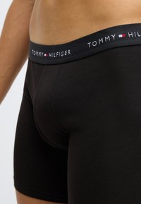 Sorte bokserbriefs lavet af glat stof med en kontrasterende elastisk talje med "TOMMY HILFIGER" logo i hvidt og rødt.