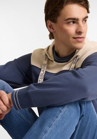 Sudadera con capucha en azul marino y beige con un acabado texturizado, que presenta detalles a rayas y cordones de marca, combinada con vaqueros celestes.