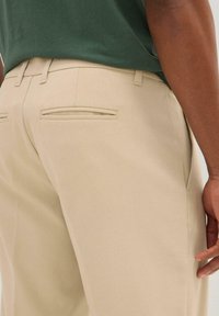 I pantaloni beige presentano un tessuto liscio, un design slim-fit e due tasche posteriori con aperture orizzontali. La cintura è piatta e confezionata.