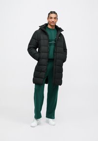 Άνδρας με πλεξούδες που φοράει μαύρο αδιάβροχο μπουφάν πάνω από πράσινη φόρμα και λευκά αθλητικά παπούτσια, στέκεται με τα χέρια στις τσέπες.