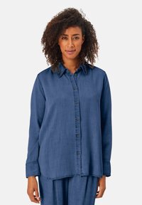 Donna con capelli ricci che indossa una camicia a maniche lunghe blu con bottoni e pantaloni larghi coordinati, davanti a uno sfondo bianco uniforme.