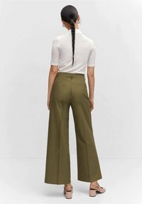 Pantalon large taille haute vert olive à la texture lisse, avec poches arrière et coupe ajustée, associé à des chaussures rose pâle.