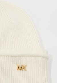 Gorro de punto color crema con textura acanalada, que presenta un dobladillo enrollado y un acento de logo metálico dorado "MK".