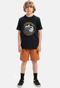 Camiseta negra de algodón con un gráfico circular que presenta olas y un sol; combinada con pantalones cortos de pana naranjas y calcetines negros con zapatos.