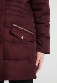 Cappotto imbottito bordeaux con un design trapuntato, dotato di una tasca con cerniera e dettagli con bottoni, realizzato in un tessuto liscio e leggero.
