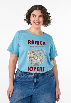 Uśmiechnięta osoba z kręconymi włosami, ubrana w niebieską koszulkę "Ramen Lovers" z motywem miski z makaronem i czerwonym napisem, do tego niebieskie dżinsy.