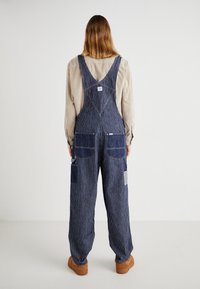 Pantalones de mezclilla con rayas verticales azules y blancas, múltiples bolsillos y un diseño en forma de V en la parte de atrás. Tejido texturizado con un ajuste relajado.