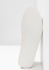 Semelle d'une basket en maille rose montrant une semelle extérieure en caoutchouc blanc texturé avec un fin motif ondulé sur fond blanc.