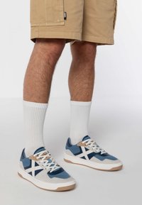 Tenisky s kombináciou modrých, sivých a bielych materiálov. Vlastnosti zahŕňajú textúrovanú látku, gumovú podrážku a svetlohnedý akcent pri podpätku.