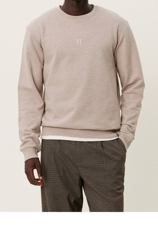 Homme portant un sweat-shirt beige clair à col rond avec un petit chiffre romain II sur la poitrine et un pantalon marron à carreaux avec la main dans la poche.