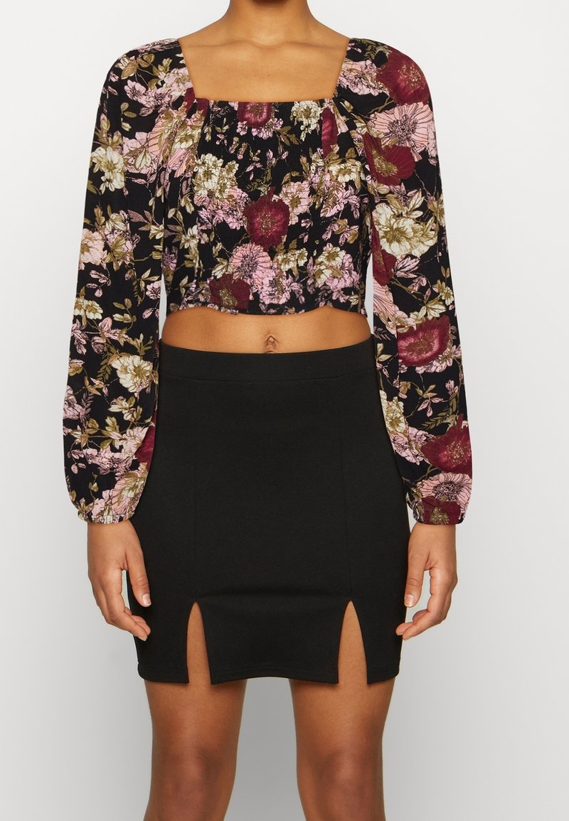 Top corto floreale con maniche lunghe in nero, caratterizzato da fiori rosa e bordeaux. Mini gonna nera a vita alta con doppie aperture laterali.