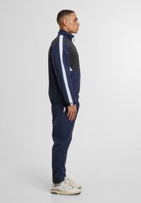 Námořnický teplákový komplet s bílými a černými akcenty, obsahující zippovou bundu, elastické manžety a odpovídající kalhoty. Hladký materiál a atletický střih.