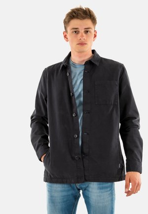 Zwarte button-up jas met een zak, gemaakt van katoenen stof. Heeft een kraag en is gecombineerd met een blauwe t-shirt en lichte jeans.