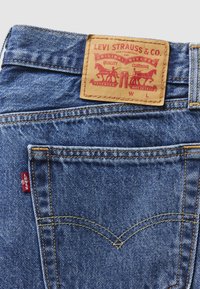 Poche arrière d'un jean Levi's bleu montrant des coutures orange, un onglet Levi's rouge et une étiquette en cuir beige avec deux chevaux et les détails de la marque.