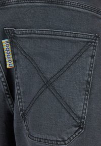 Tissu en denim noir avec une poche arrière visible, présentant des coutures contrastées et une étiquette bleu et jaune sur laquelle est écrit "homeboy."