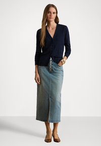 Tmavomodrý kardigán so zapínaním na gombíky, kombinovaný so svetlomodrou džínsovou maxi sukňou s predným rozparkom a veľkými gombíkmi, nosený s hnedými lodičkami.