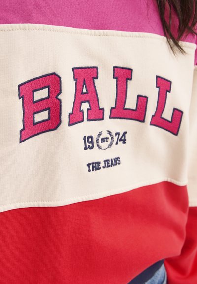 Krāsains džemperis ar drosmīgiem sarkaniem, rozā un krēmkrāsas svītrām. Teksts saka "BOLA" lielā fontā ar izšūtiem sīktēliem un apaļu logo.