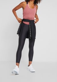 Leggings de sport noirs associées à un débardeur bordeaux, surmonté d'une veste noire nouée à la taille. Tissus lisses et baskets blanches.