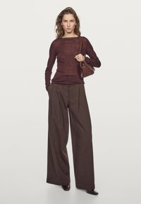 Ciemnobrązowe szerokie spodnie połączone z dopasowaną, przezroczystą bordową bluzką z długim rękawem. Modelka trzyma małą brązową torebkę. Gładkie faktury.
