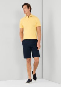 Žluté polo tričko s límečkem, spárované s námořnickými šortkami a tmavými nazouvacími botami. Tkanina vypadá hladce a lehce. Jednoduchý design.