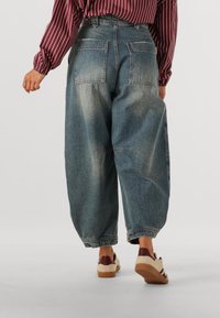 Baggy denim jeans in een vervaagde blauwe kleur, met twee achterzakken en een iets taps toelopende pijp, gecombineerd met bruin en crèmekleurige sneakers.