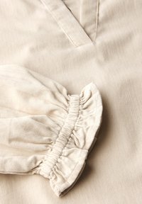 Beige Cord-Stoff mit gerafftem, elastischem Bündchen und überlappender V-förmiger Ausschnittnaht.