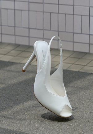 Chaussure blanche à talon haut avec bride arrière et bout ouvert sur le trottoir devant un mur carrelé, projetant une ombre sur le sol.