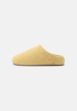 Chausson jaune doux à enfiler, bout fermé, petit talon, texture en daim et logo UGG embossé près du talon sur fond blanc.