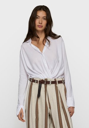 Kvinde med langt mørkt hår iført hvid skjorte med knapper, stoppet ned i beige bukser med lodrette brune striber og et mørkebrunt bælte.