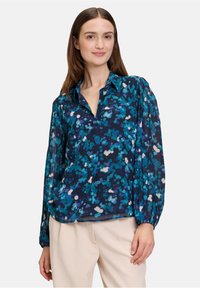 Bloemige blouse in donkerblauw met grote teal- en witte patronen. Voorzien van een kraag, knoopsluiting aan de voorkant en lange, licht opgeblazen mouwen.