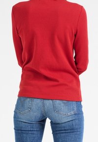 Personne portant une chemise rouge à manches longues en coton côtelé et un jean en denim bleu, vue de dos sur un fond blanc uni.
