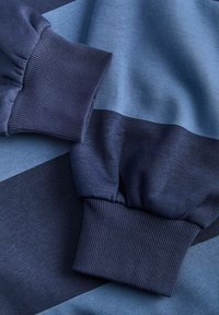 Zils un tumši zils džemperis ar ribotām piedurknēm, gludu tekstūru un plašām horizontālām svītrām dažādos zilās toņos.