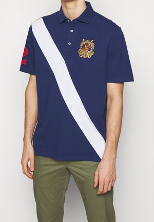 Uomo che indossa una polo blu navy con una striscia diagonale bianca, stemma ricamato sul petto, numero 2 rosso sulla manica e pantaloni verde oliva.