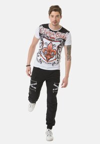 Weißes T-Shirt mit orangefarbenem und schwarzem grafischem Design, schwarze Cargo-Hosen mit Reißverschlüssen und weißem Text, weiße Sneakers, männliches Model in Bewegung.