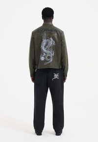 Giacca di pelle verde oliva con una grande grafica di un drago grigio sulla schiena, abbinata a pantaloni neri larghi con un logo bianco sul retro.