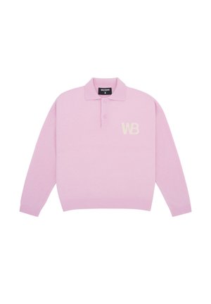 Femme aux cheveux bouclés et foncés portant un pull polo rose clair à manches longues, un jean large bleu clair et des chaussures noires, debout devant un fond blanc.