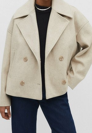 Veste beige en laine à double boutonnage avec larges revers, portée sur un pull noir et un jean bleu foncé, accessoirisée d'un collier argenté.