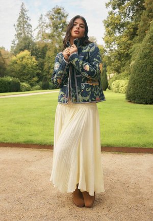 Femme portant une veste matelassée à fleurs, une jupe plissée crème et des bottines brunes, debout sur un chemin de jardin avec de la verdure en arrière-plan.