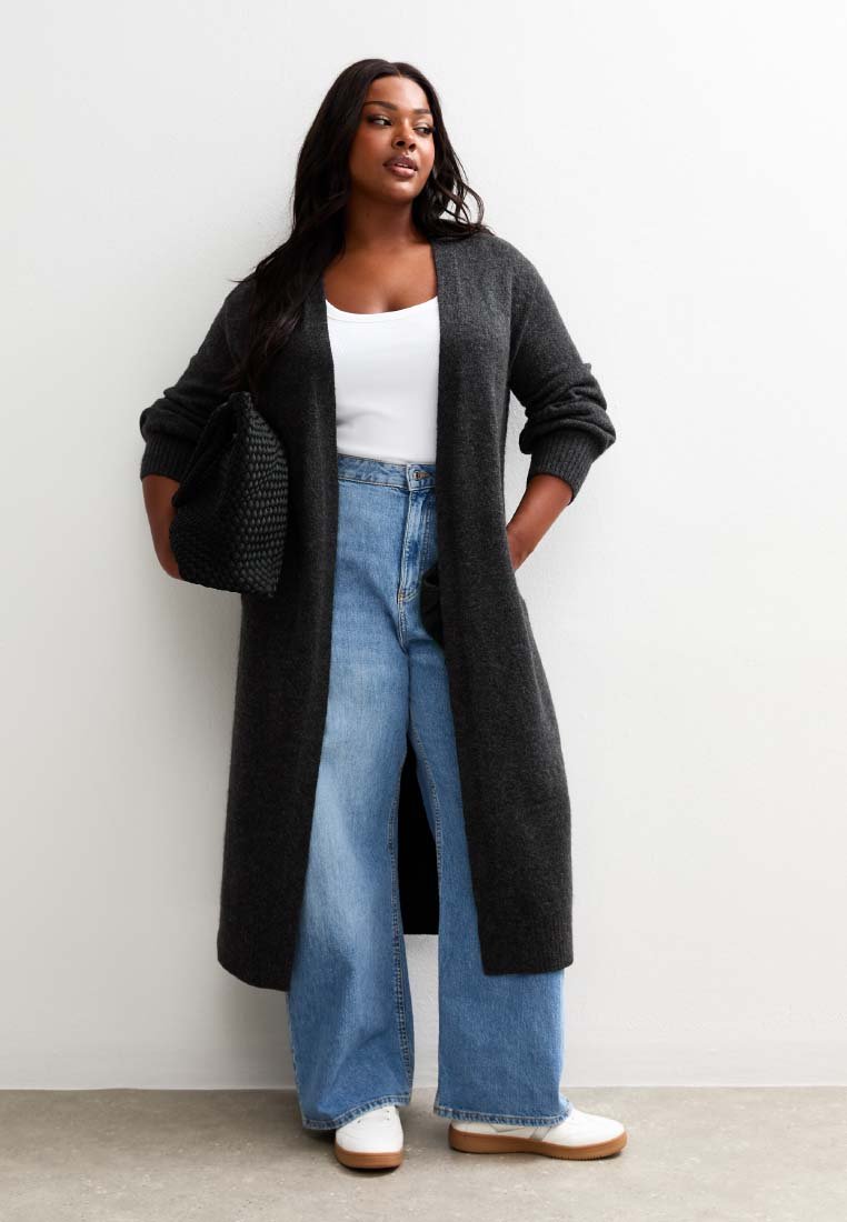 Vrouw in een lange donkergrijze cardigan, witte top, wijde blauwe jeans, witte schoenen, met een gestructureerde zwarte tas, staand tegen een witte muur.