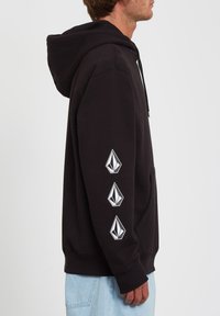 Felpa nera con cappuccio e tasca frontale, con cordoncino per il cappuccio e tre accenti bianchi del logo disposti verticalmente sulla manica sinistra. Tessuto morbido con vestibilità comoda.