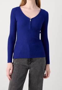 Haut à manches longues côtelé de couleur bleu vif avec un col en V et un petit zip à l'avant, associé à un pantalon slim gris foncé.