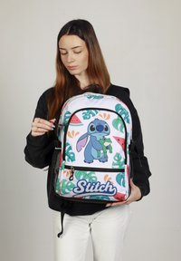 Mochila con un gráfico colorido de Stitch, fabricada de tela blanca y con patrones de hojas tropicales verdes. Cuenta con compartimentos con cremallera y laterales de malla.