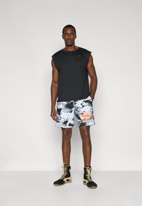 Chemise noire sans manches, short de bain à motifs multicolores avec des accents gris et orange, et chaussures noires avec des lacets jaunes.