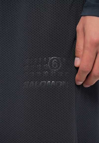 Juodas tekstūruotas audinys su šešiakampiu raštu, kuriame yra iškilūs skaičiai ir „SALOMON“ logotipas šalia apvado, ranka laikanti drabužį.
