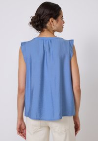 Top blu senza maniche con una finitura testurizzata, dettaglio arricciato sul retro e leggero volant sulle spalle. Abbinato a pantaloni beige.